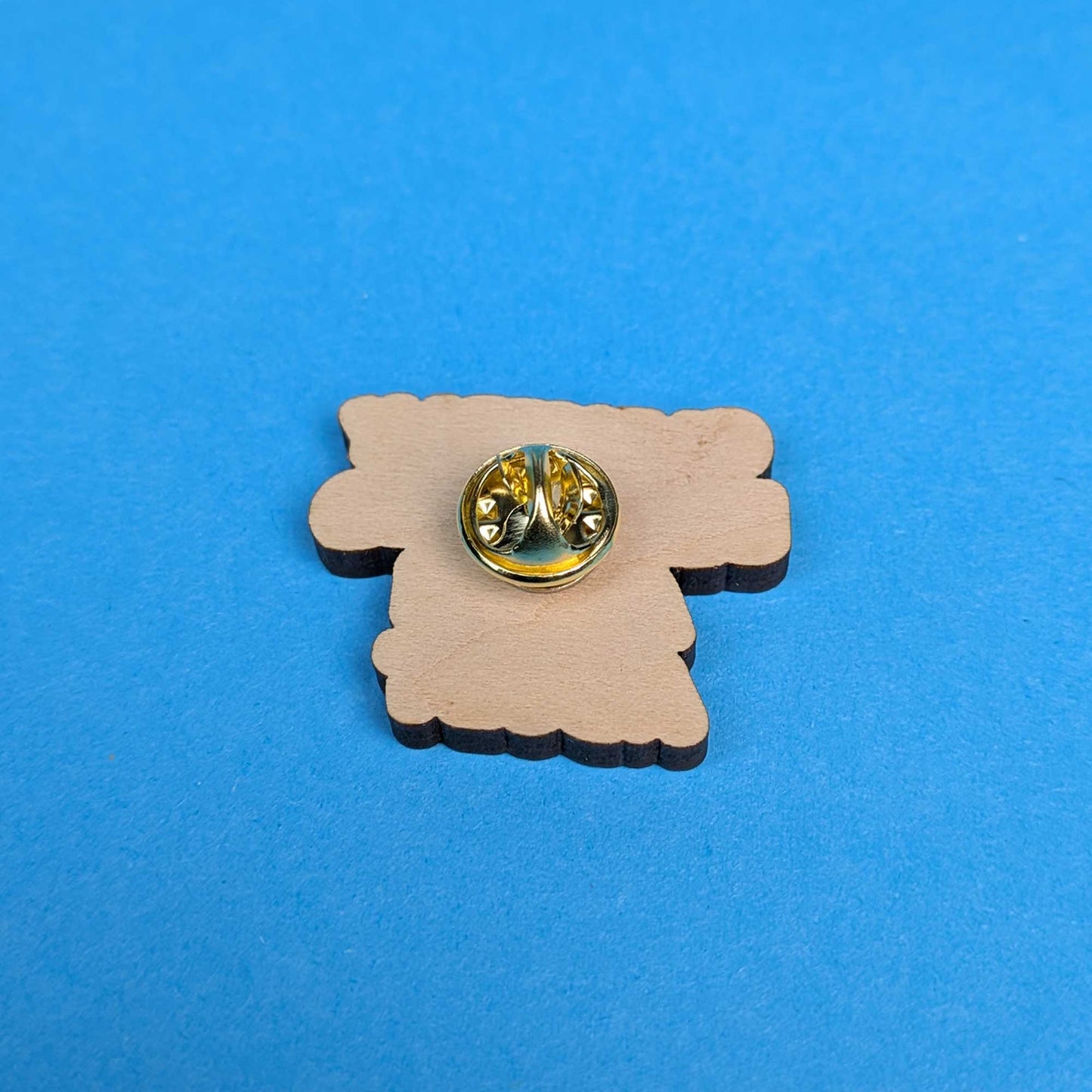 A small wooden charm or badge with a scalloped edge, featuring a gold-tone metal pin back, placed on a solid blue background.