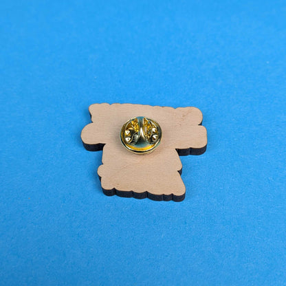 A small wooden charm or badge with a scalloped edge, featuring a gold-tone metal pin back, placed on a solid blue background.