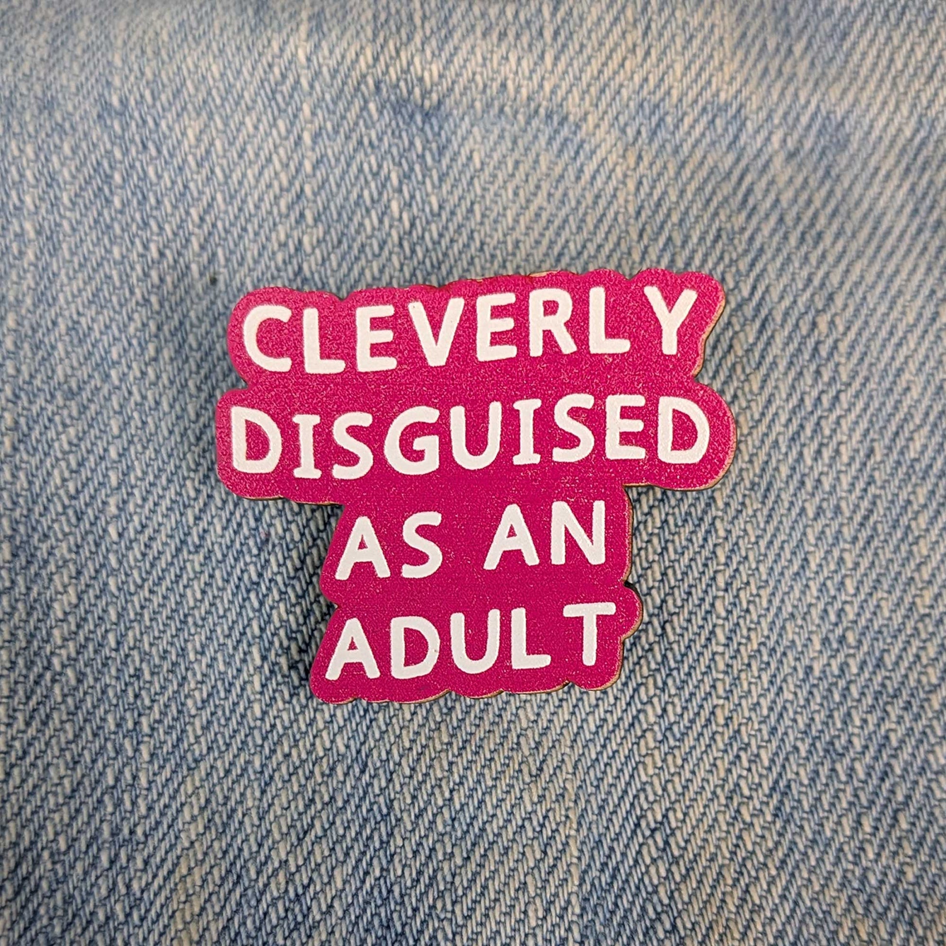 A bright pink enamel pin with white text reading 'CLEVERLY DISGUISED AS AN ADULT' placed on a blue denim background.