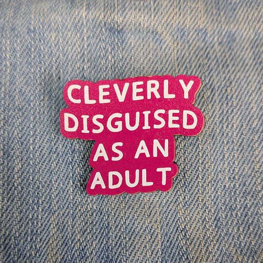 A bright pink enamel pin with white text reading 'CLEVERLY DISGUISED AS AN ADULT' placed on a blue denim background.