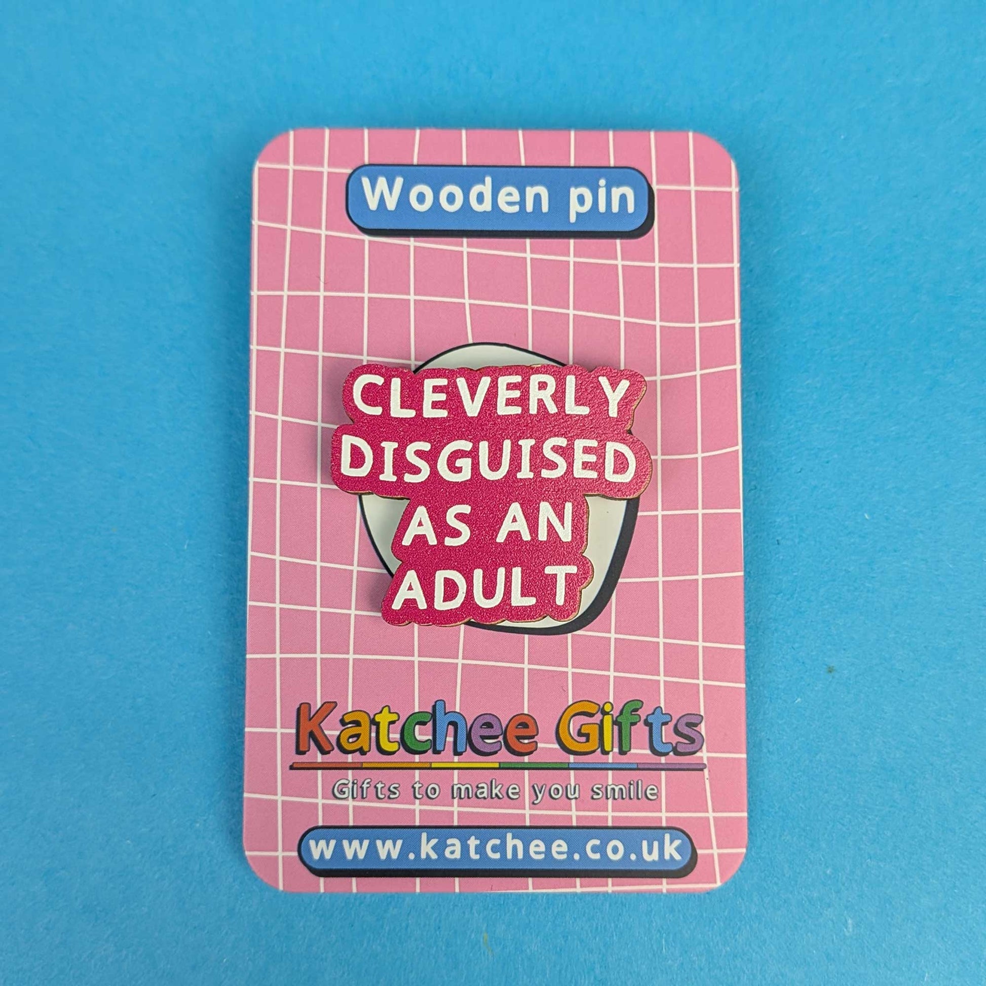A wooden pin displayed on its backing card, featuring the text 'CLEVERLY DISGUISED AS AN ADULT' in white lettering on a pink and red background. The backing card is pink with a white grid pattern and displays the brand 'Katchee Gifts' at the bottom, set against a solid blue background.