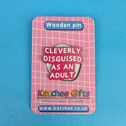 A wooden pin displayed on its backing card, featuring the text 'CLEVERLY DISGUISED AS AN ADULT' in white lettering on a pink and red background. The backing card is pink with a white grid pattern and displays the brand 'Katchee Gifts' at the bottom, set against a solid blue background.