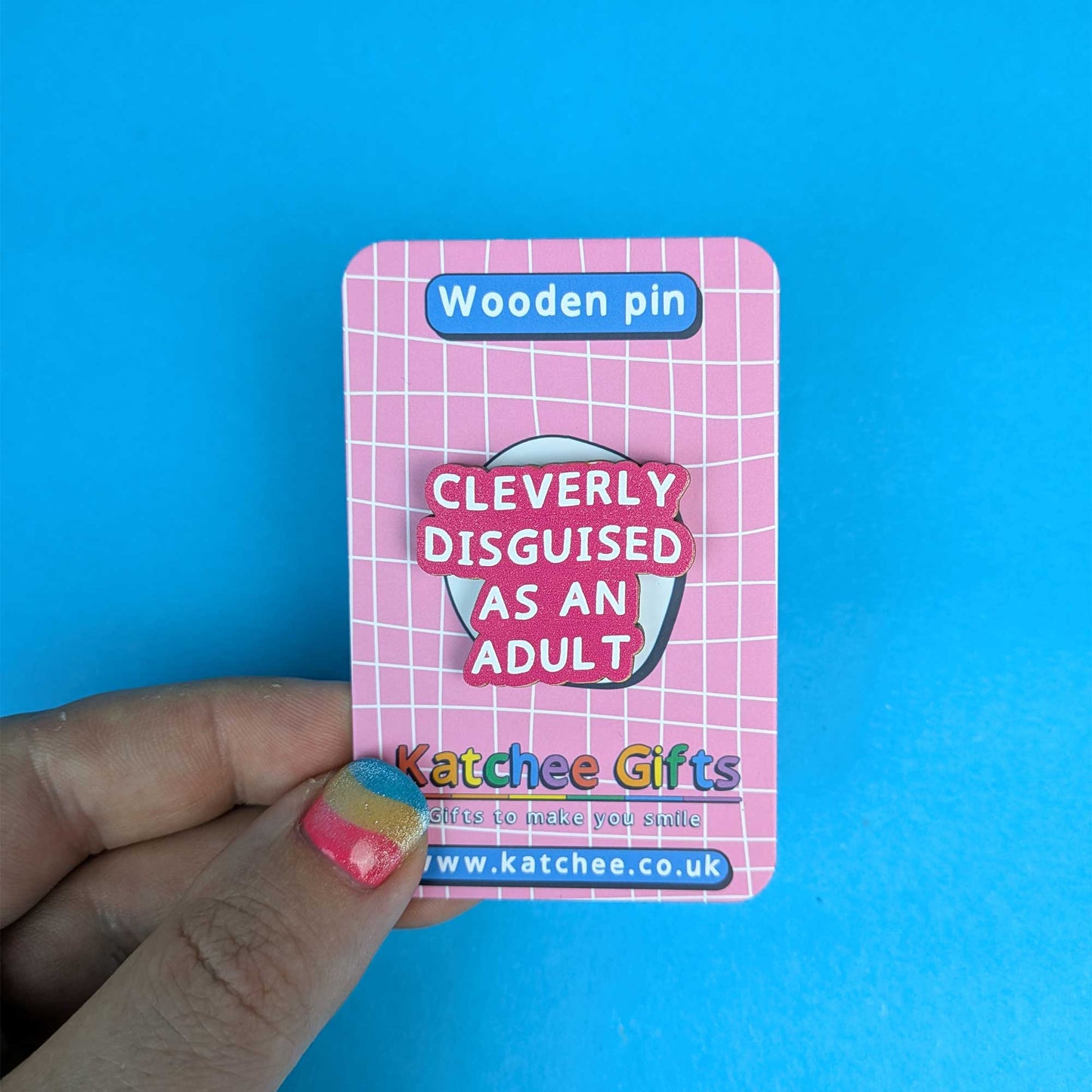 A hand holds a pink card backing featuring a wooden pin that reads 'Cleverly Disguised As An Adult' against a bright blue background.