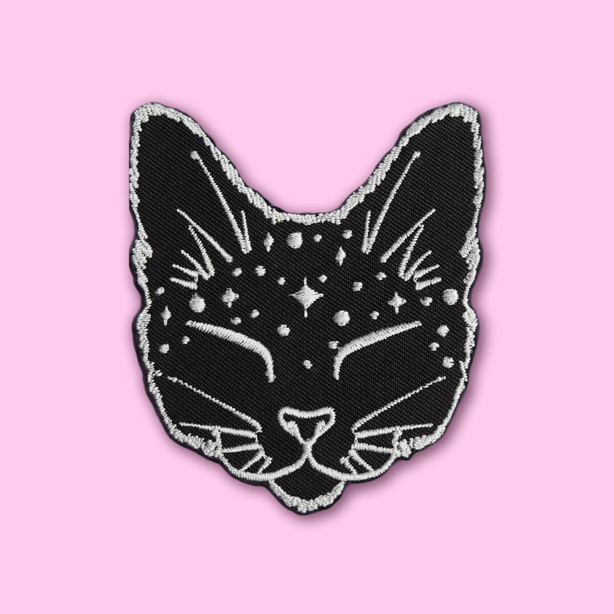 A black cat head embroidered patch with white star and line details, outlined in white, shown on a solid pink background.