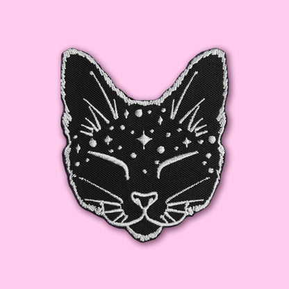 A black cat head embroidered patch with white star and line details, outlined in white, shown on a solid pink background.