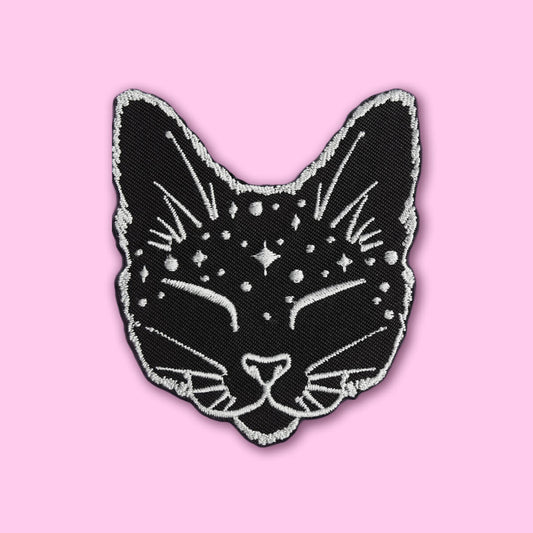A black cat head embroidered patch with white star and line details, outlined in white, shown on a solid pink background.