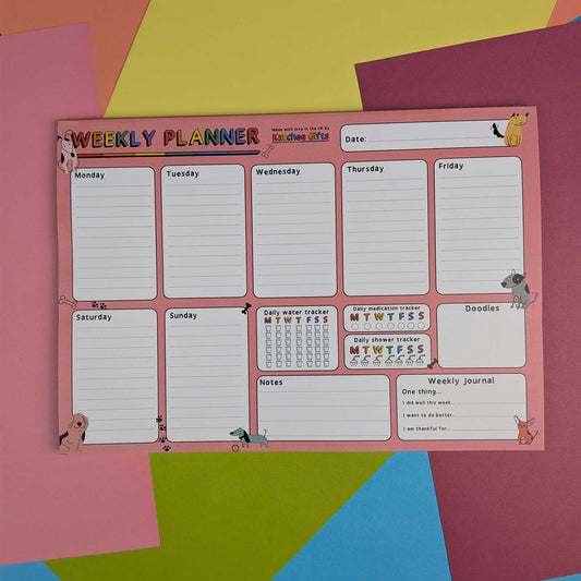 A pink weekly planner board with lined sections for Monday through Sunday, a notes area, and a small daily water tracker, photographed on a colorful geometric paper background.