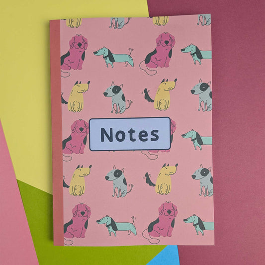 A pink hardcover notebook with a repeating pattern of cartoon dachshunds in pink, yellow, and blue-gray, featuring a light blue label in the center that reads 'Notes'. The notebook is photographed on a colorful background with pink, yellow, green, and blue paper shapes.