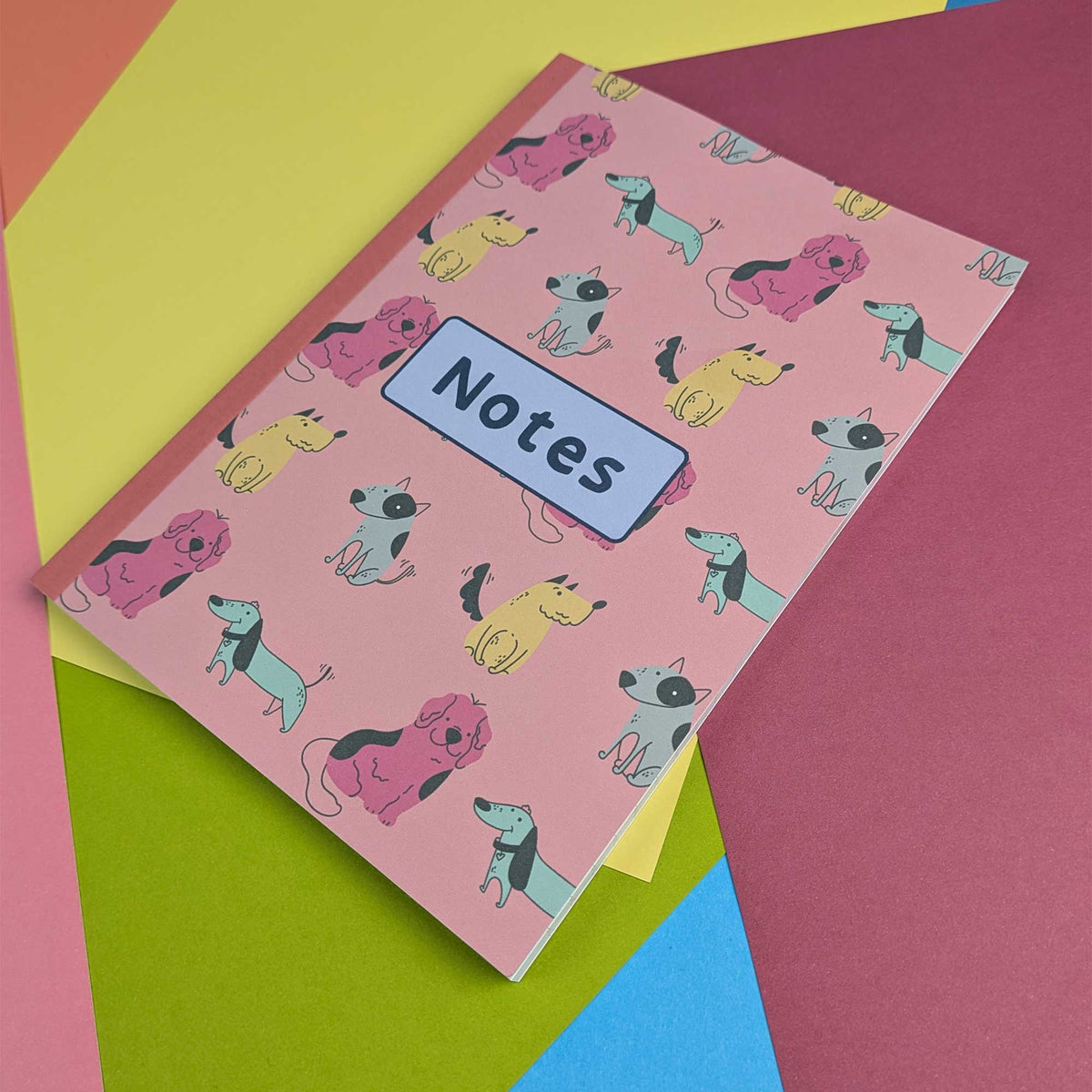 A pink hardcover notebook with a repeating pattern of cartoon dachshunds in pink, yellow, blue, and black, featuring a light blue label that says 'Notes'. The notebook is styled on top of a stack of colorful paper sheets in yellow, green, magenta, and blue.