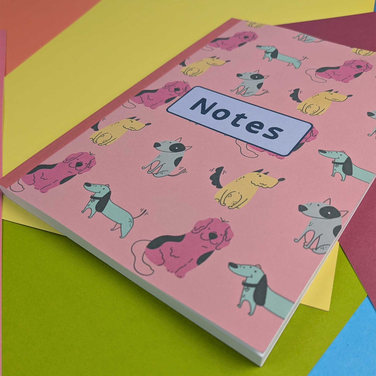 A pink hardcover notebook with a cartoon dog pattern and a white label reading 'Notes' on the cover, photographed on top of colorful paper sheets.