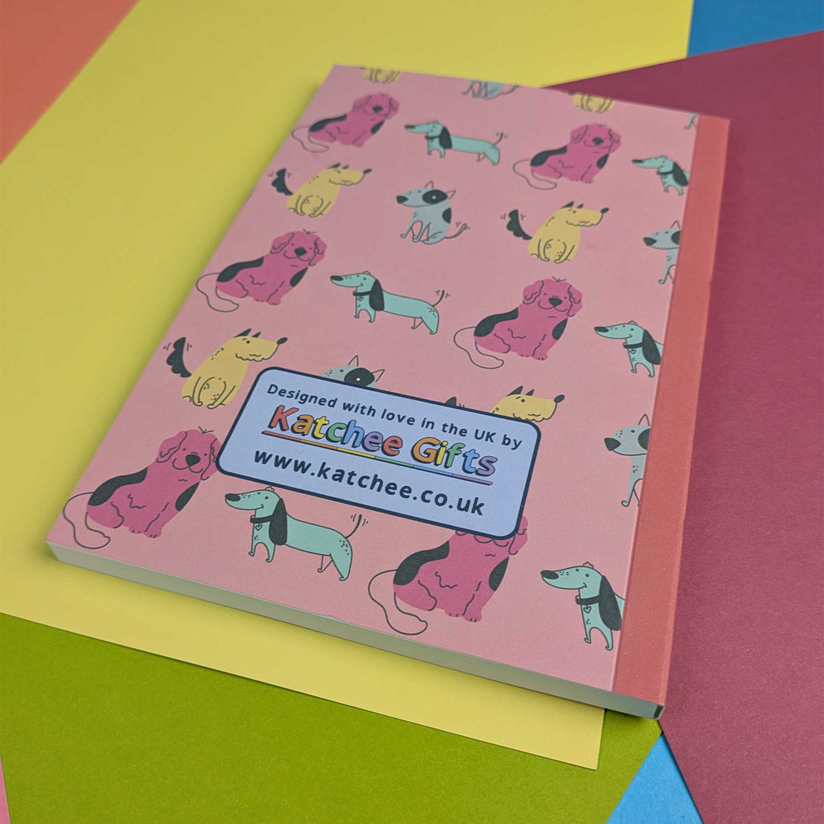 A pink hardcover notebook featuring a colorful pattern of cartoon dogs, photographed on top of layered pastel paper sheets. A label on the cover reads 'Designed with love in the UK by Katchee Gifts www.