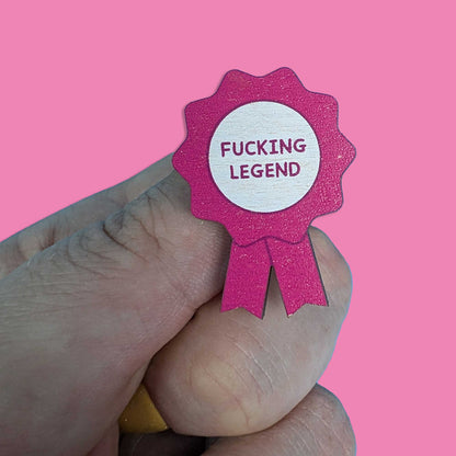 A hand holding a small pink ribbon badge with a glittery texture and a white center that reads 'FUCKING LEGEND' in pink text, set against a solid pink background.