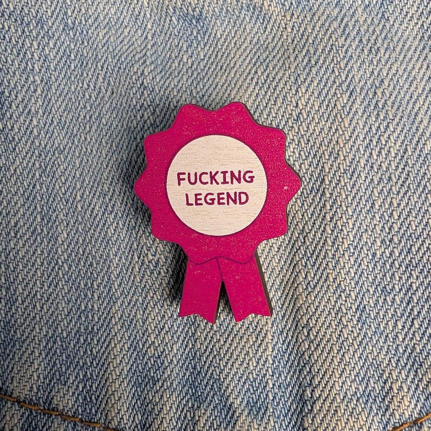 A small pink ribbon-shaped pin or badge with a light wood center reading “FUCKING LEGEND” in pink text, placed on blue denim fabric.