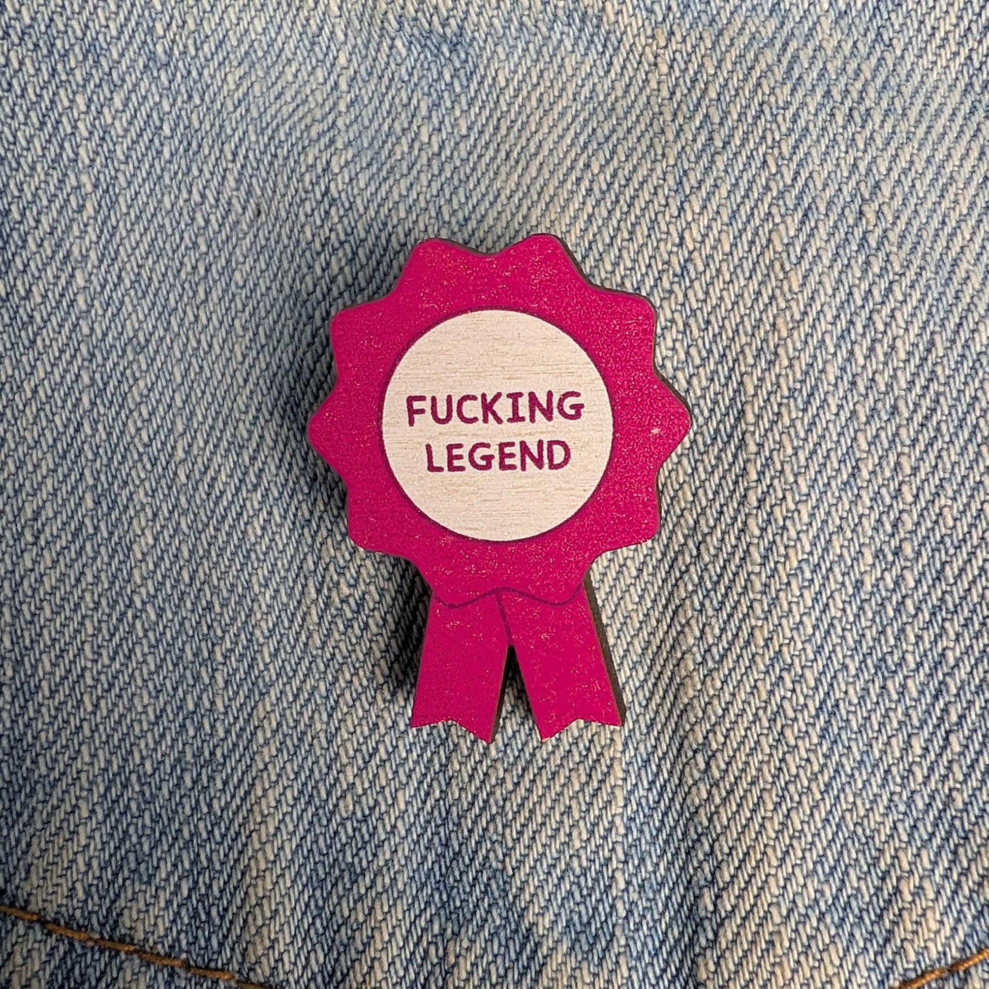 A small pink ribbon-shaped pin or badge with a light wood center reading “FUCKING LEGEND” in pink text, placed on blue denim fabric.