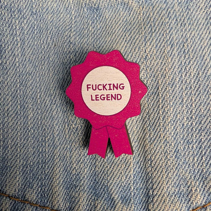 A small pink ribbon-shaped pin or badge with a light wood center reading “FUCKING LEGEND” in pink text, placed on blue denim fabric.