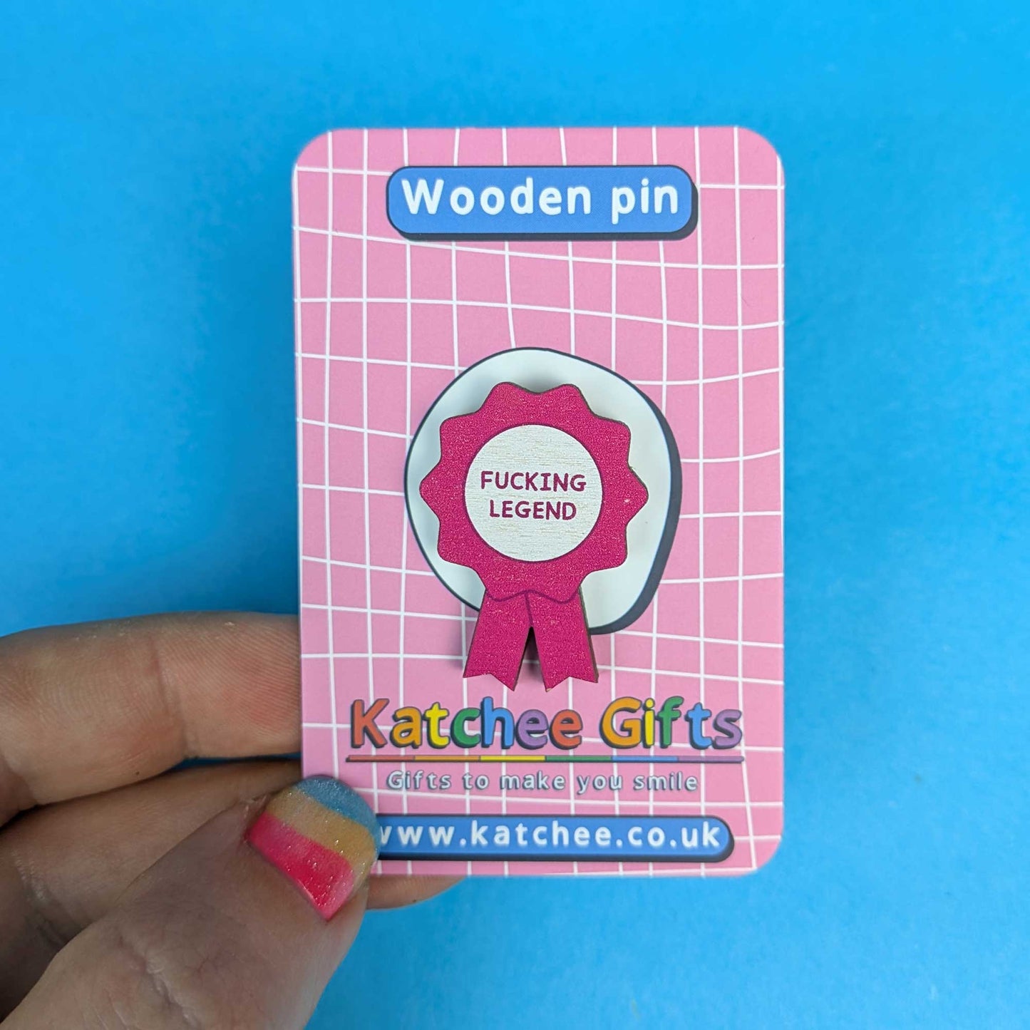 A hand holds a pink card featuring a wooden pin. The pin is a ribbon-style badge with a white center and a pink scalloped border, reading 'FUCKING LEGEND'.