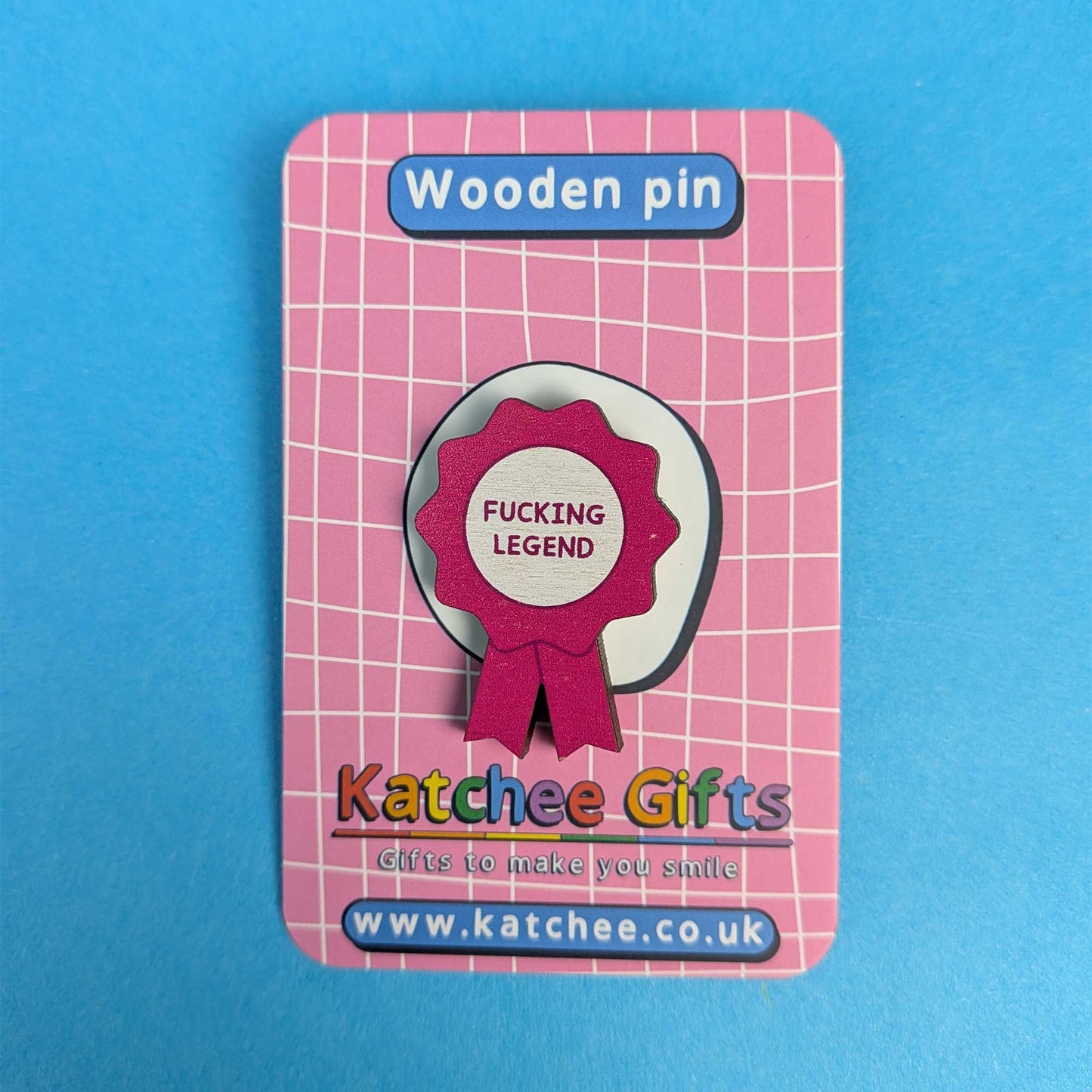 A wooden pin badge displayed on its pink branded backing card. The pin is a circular wooden badge with a magenta ribbon seal that reads 'FUCKING LEGEND'.