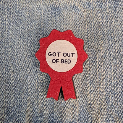 A red ribbon-shaped wooden pin with a white center reading 'GOT OUT OF BED' in black text, placed on a blue denim fabric background.