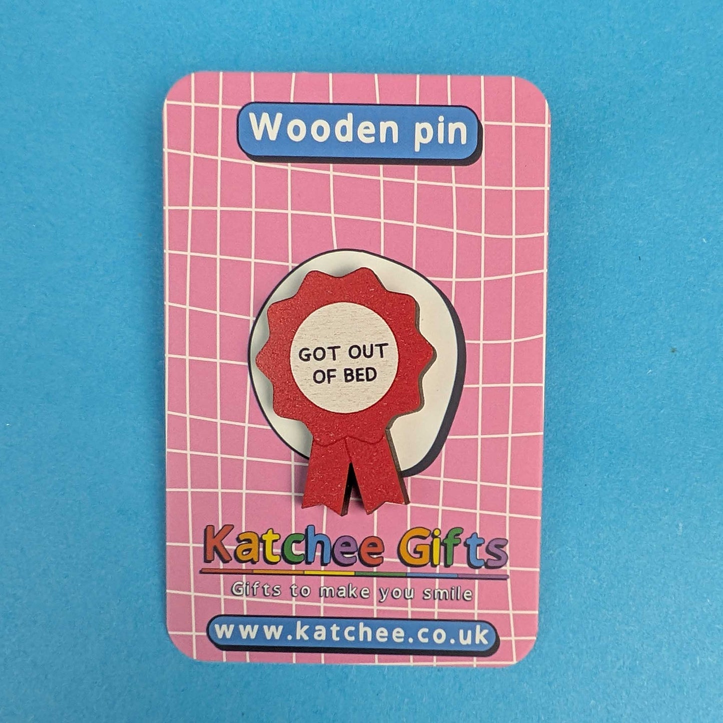 A wooden pin shaped like a red ribbon badge with the text 'GOT OUT OF BED' on a pink card backing. The card shows the brand name 'Katchee Gifts' and website at the bottom, set against a solid blue background.