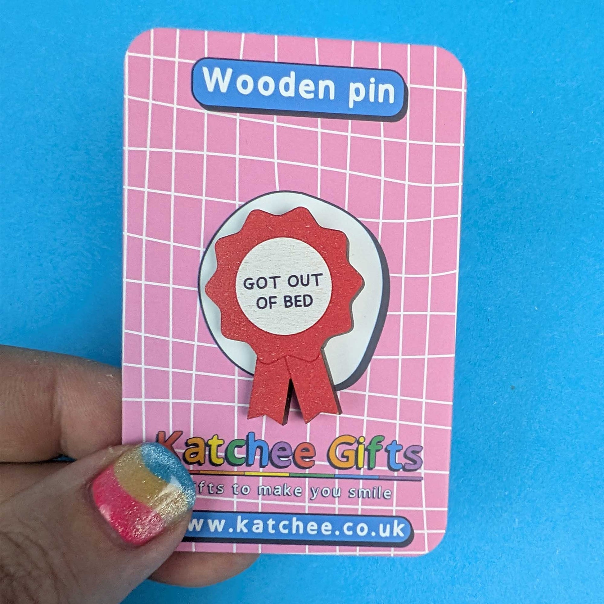 A wooden pin badge shaped like a red ribbon seal with the text 'GOT OUT OF BED' on a pink card backing, held by a hand against a bright blue background. The card is branded 'Katchee Gifts'.