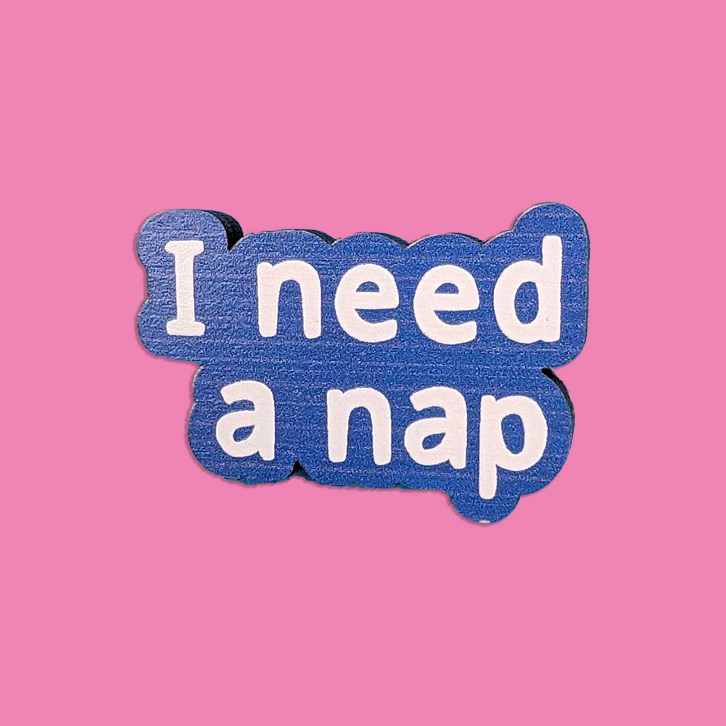 A die-cut wooden pin with the text 'I need a nap' in white lettering on a textured blue wood background, set against a solid pink backdrop.