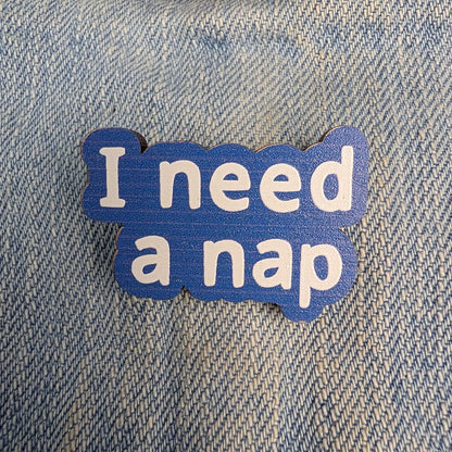A blue enamel lapel pin with the text 'I need a nap' in white lowercase letters, displayed on a blue denim fabric background.