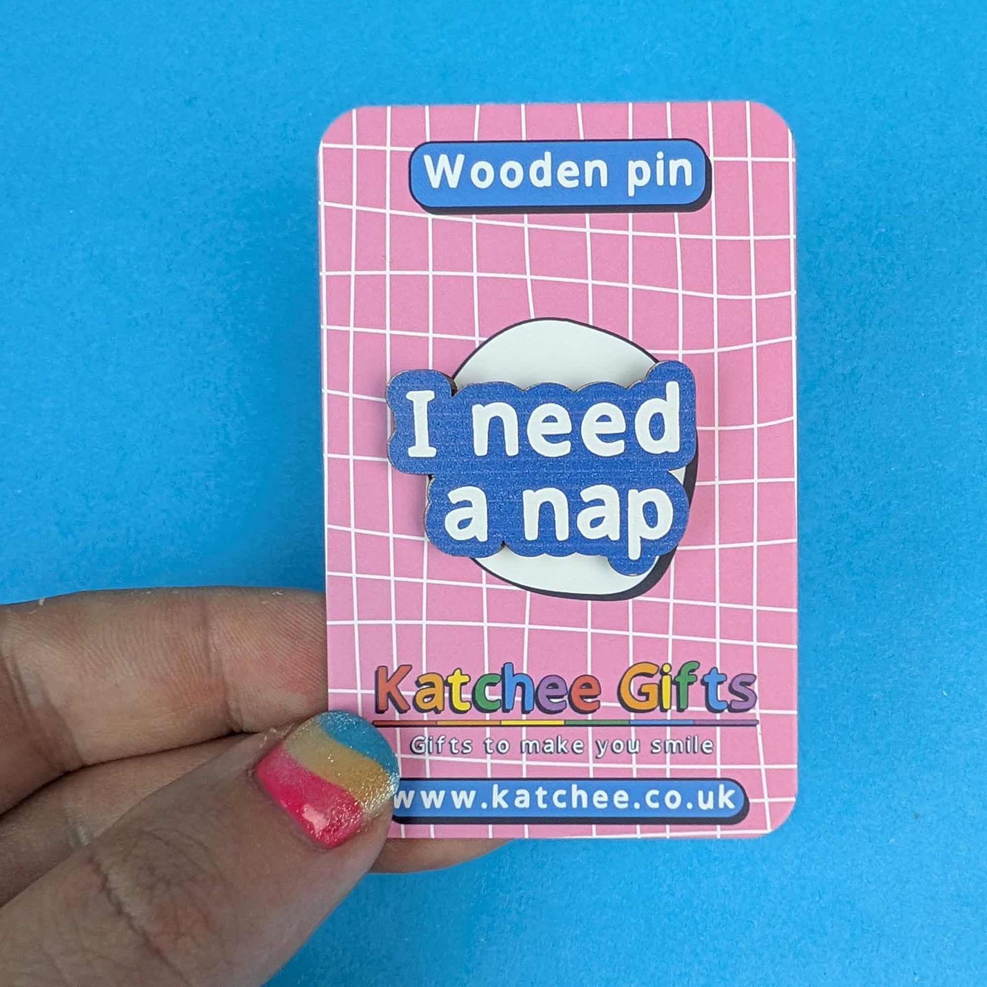 A hand holds a pink card backing for a wooden pin that reads 'I need a nap' in blue and white lettering. The card says 'Wooden pin' at the top and shows the brand 'Katchee Gifts' with website text at the bottom, against a bright blue background.