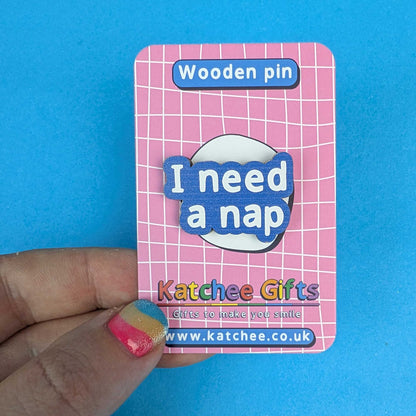 A hand holds a pink card backing for a wooden pin that reads 'I need a nap' in blue and white lettering. The card says 'Wooden pin' at the top and shows the brand 'Katchee Gifts' with website text at the bottom, against a bright blue background.