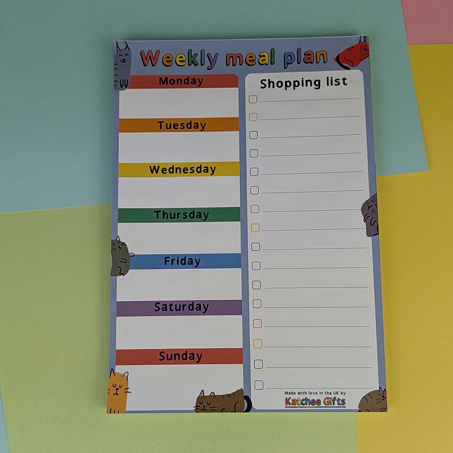 Weekly meal plan and shopping list notepad with colorful days of the week labels on a multicolored background.
