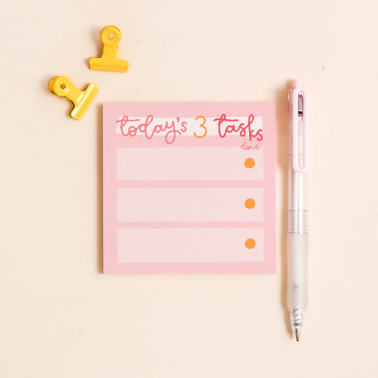 Pink notepad with 'today's 3 tasks' text, yellow clips, and a pen on a light beige background