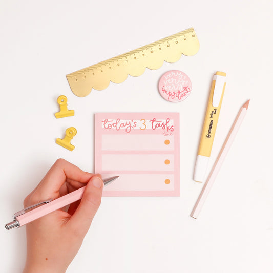 Hand holding a pen with a pink notepad labeled 'Today's 3 Tasks' on a white background.