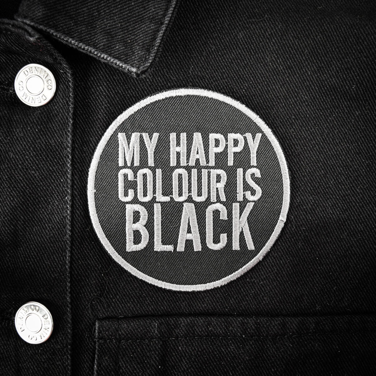 A circular embroidered patch with the text 'MY HAPPY COLOUR IS BLACK' in white on a black denim jacket, featuring silver buttons on the left.