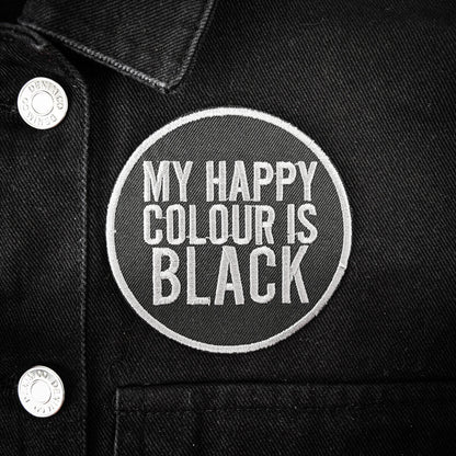 A circular embroidered patch with the text 'MY HAPPY COLOUR IS BLACK' in white on a black denim jacket, featuring silver buttons on the left.
