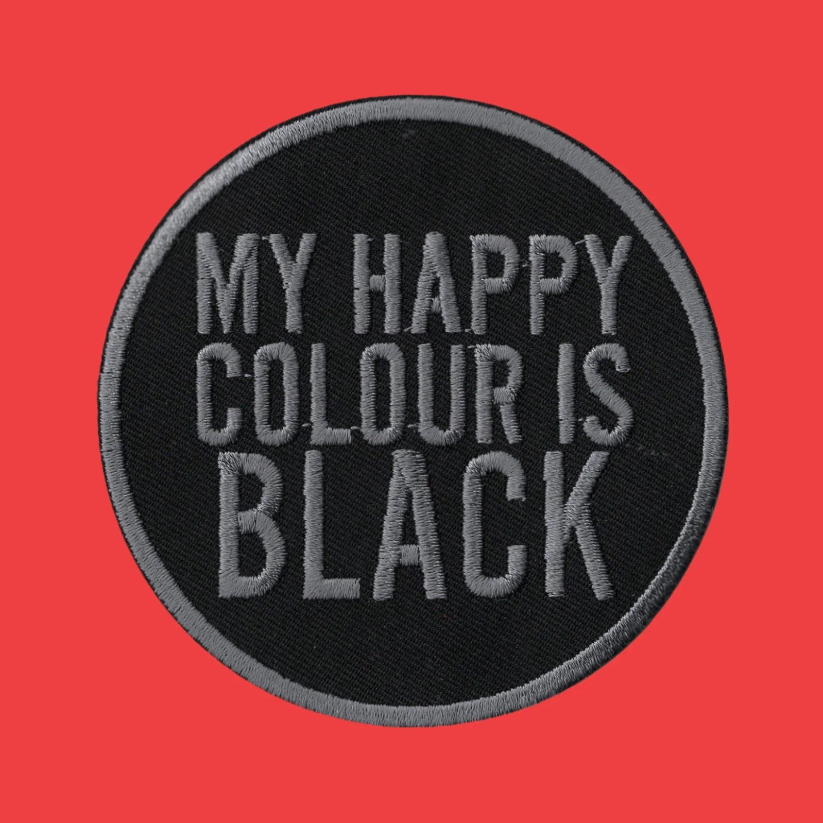 A circular embroidered patch with a black background and gray text reading 'MY HAPPY COLOUR IS BLACK', shown on a solid red background.