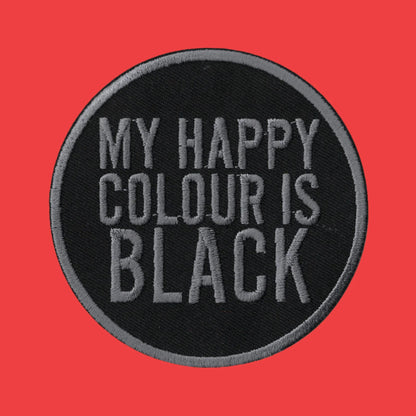 A circular embroidered patch with a black background and gray text reading 'MY HAPPY COLOUR IS BLACK', shown on a solid red background.