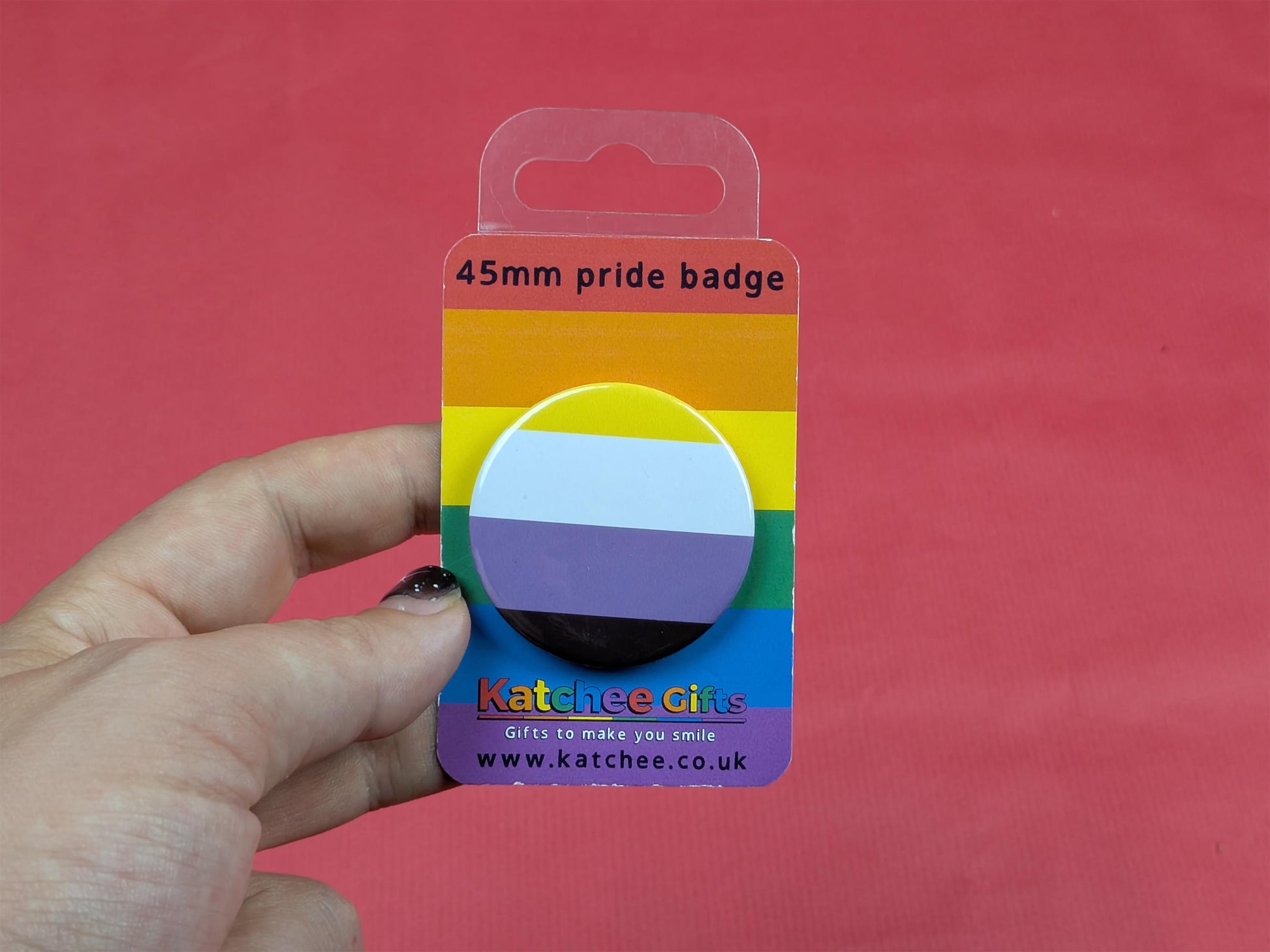 Hand holding a non binary flag 45mm pride badge with 'Katchee Gifts' packaging against a red background.