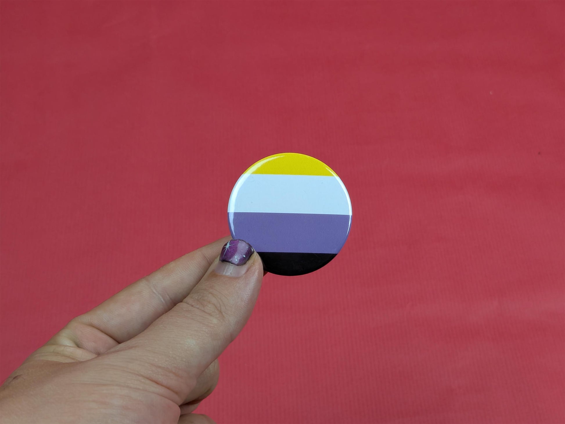 Hand holding a circular badge with a non binary flag design against a red background