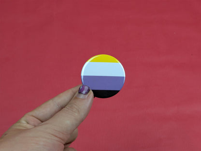 Hand holding a circular badge with a non binary flag design against a red background