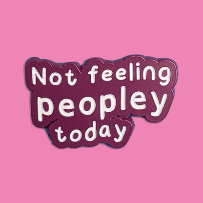 A die-cut enamel pin with the text 'Not feeling peopley today' in white lowercase letters on a dark plum purple background, set against a solid pink backdrop.