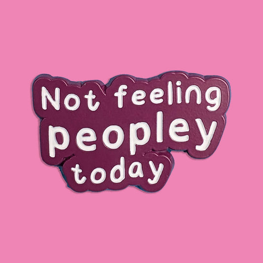 A die-cut enamel pin with the text 'Not feeling peopley today' in white lowercase letters on a dark plum purple background, set against a solid pink backdrop.