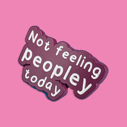 A die-cut enamel pin with the text 'Not feeling peopley today' in white lettering on a dark plum-purple background, set against a solid pink backdrop.