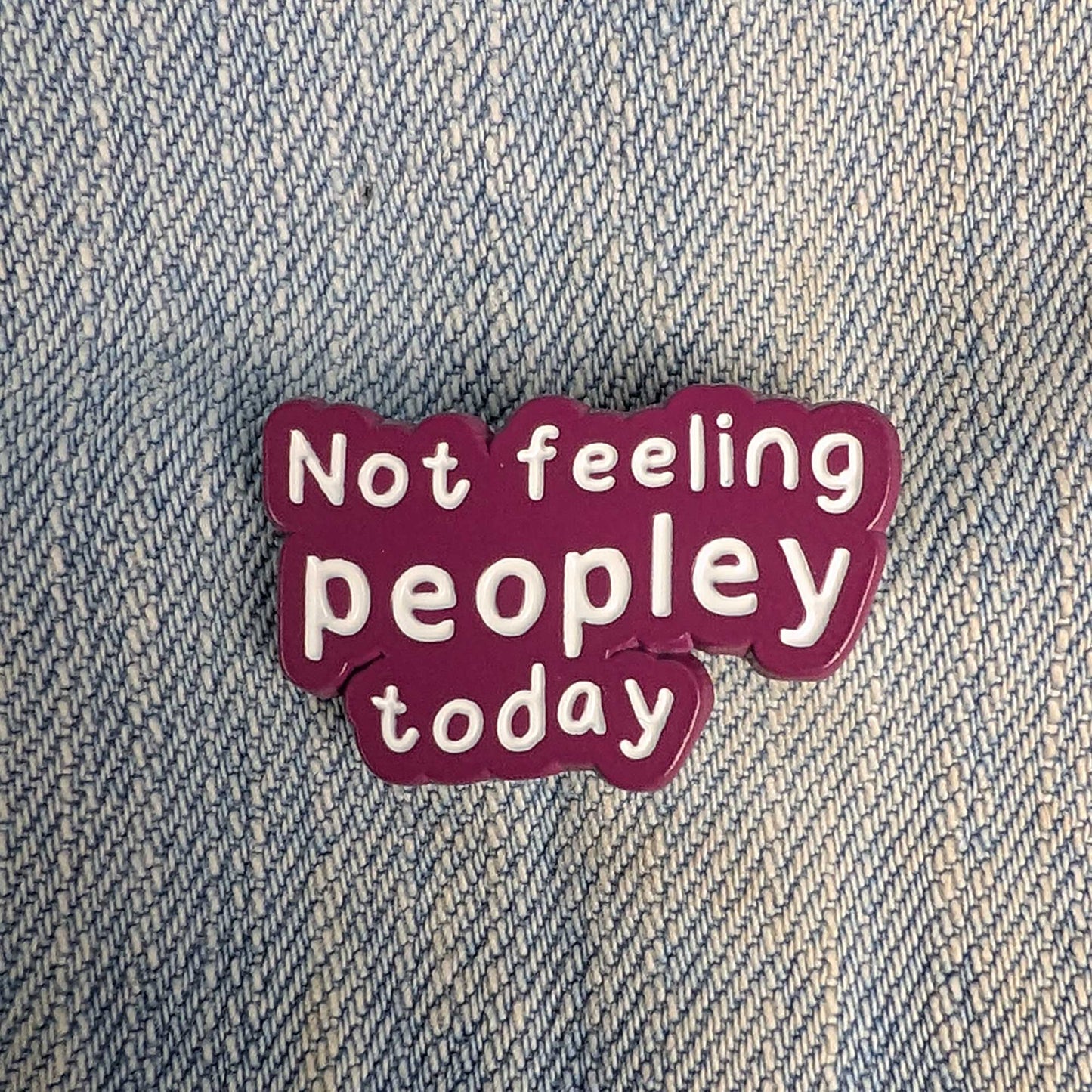 A small enamel pin with the text 'Not feeling peopley today' in white lettering on a dark magenta background, placed on a blue denim fabric surface.