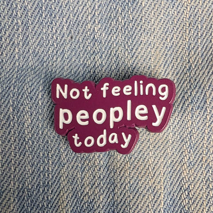 A small enamel pin with the text 'Not feeling peopley today' in white lettering on a dark magenta background, placed on a blue denim fabric surface.