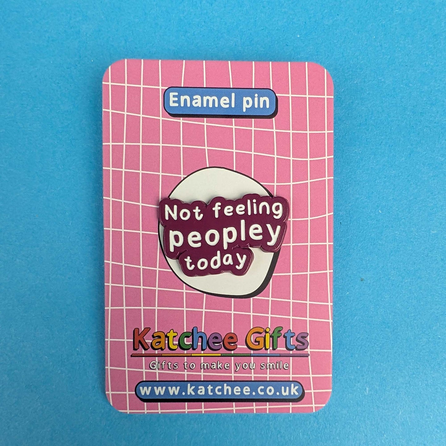 A pink enamel pin backing card featuring a white grid pattern, with a central oval design that reads 'Not feeling peopley today' in white text on a dark red background. The card is labeled 'Enamel pin' at the top and shows the brand 'Katchee Gifts' with website details at the bottom, set against a solid blue background.