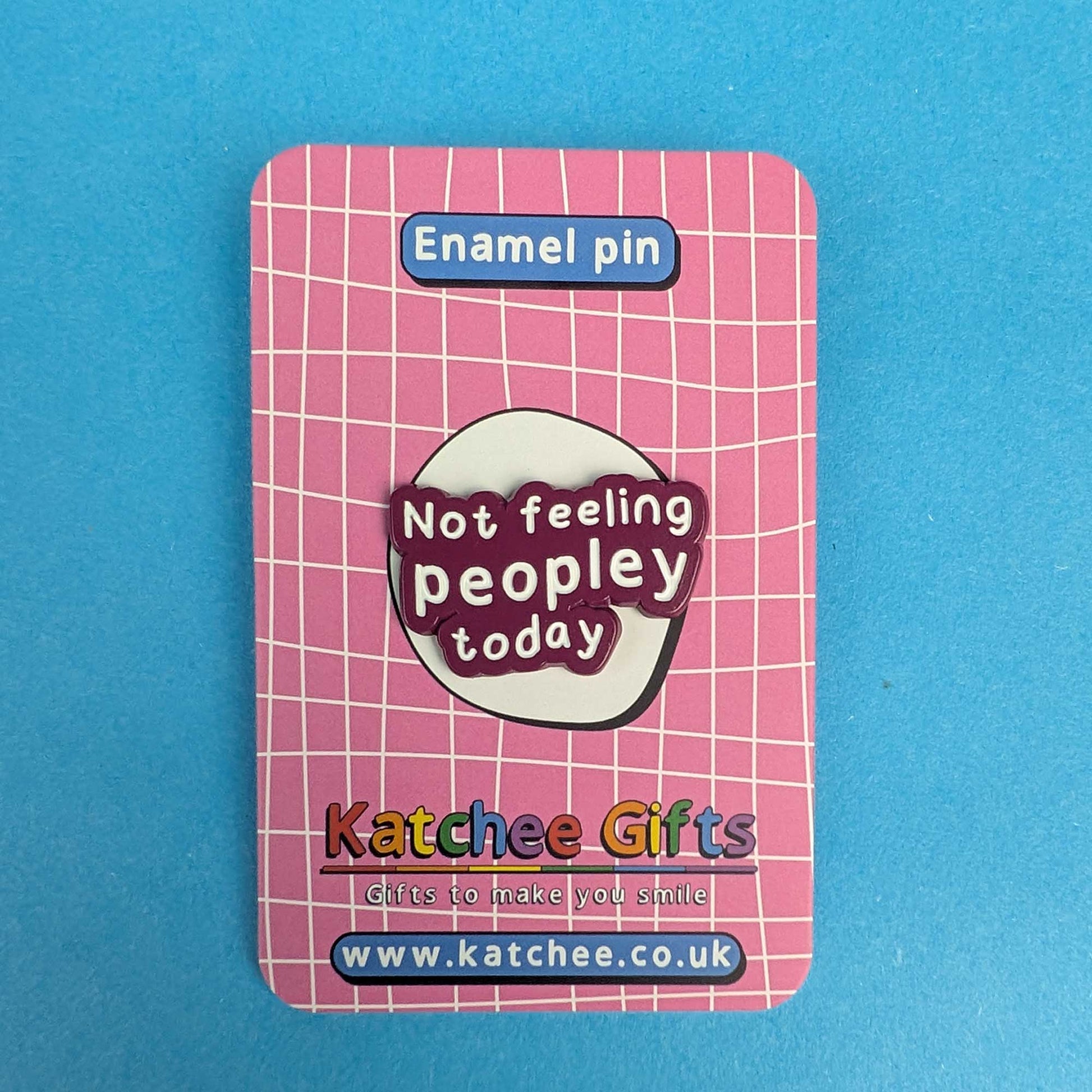 A pink enamel pin backing card featuring a white grid pattern, with a central oval design that reads 'Not feeling peopley today' in white text on a dark red background. The card is labeled 'Enamel pin' at the top and shows the brand 'Katchee Gifts' with website details at the bottom, set against a solid blue background.