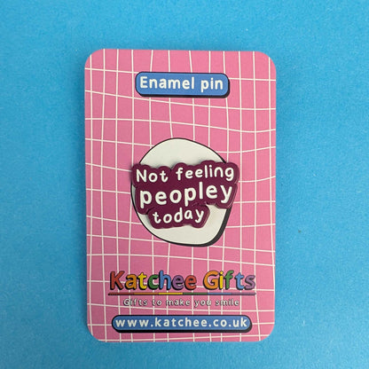 A pink enamel pin backing card featuring a white grid pattern, with a central oval design that reads 'Not feeling peopley today' in white text on a dark red background. The card is labeled 'Enamel pin' at the top and shows the brand 'Katchee Gifts' with website details at the bottom, set against a solid blue background.