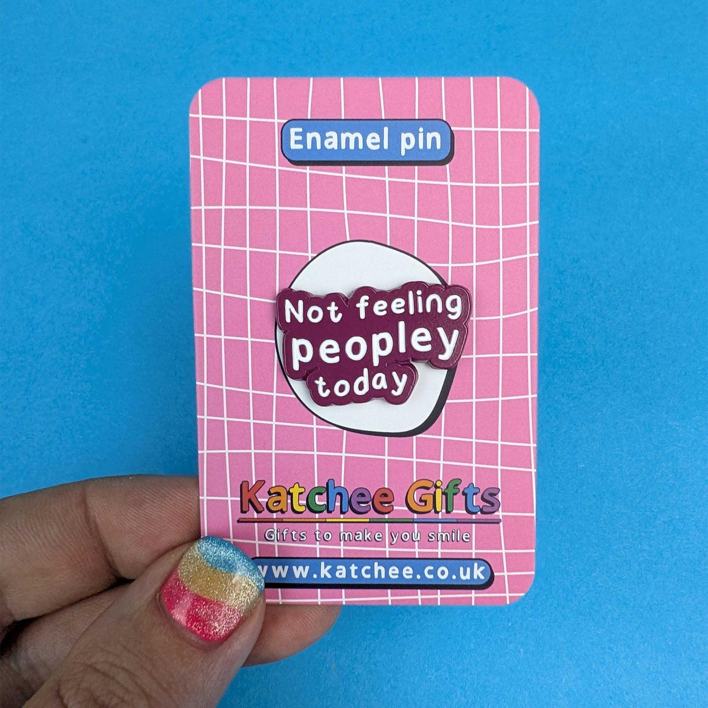 A hand holds a pink enamel pin backing card featuring a white grid pattern and a central sticker that reads 'Not feeling peopley today' in white text on a dark purple badge. The card also shows the brand name 'Katchee Gifts' and website URL at the bottom, set against a solid blue background.