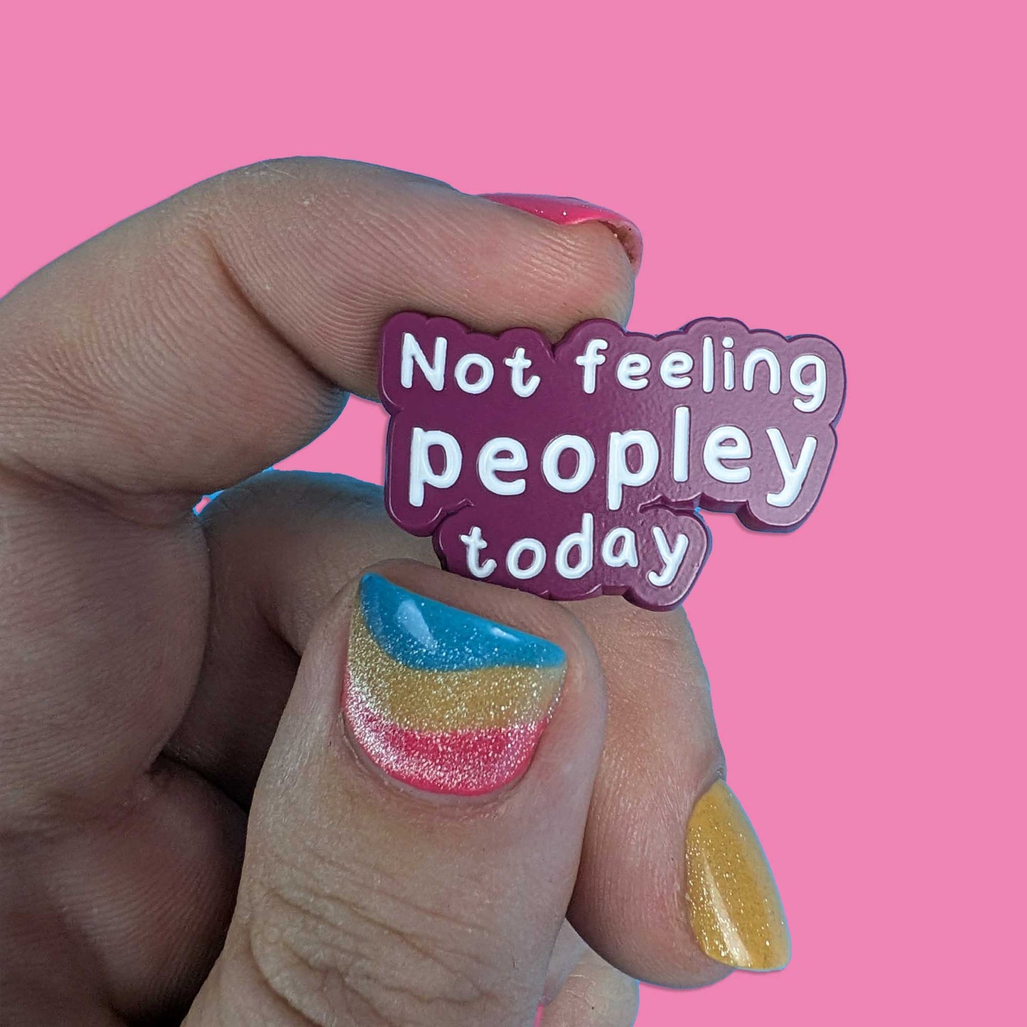 A hand with colorful glittery nail polish holds a small enamel pin that reads 'Not feeling peopley today' in white text on a purple and pink background, set against a solid pink backdrop.