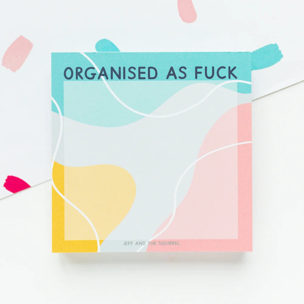A square sticky note pad with a colorful abstract design and the text 'ORGANISED AS FUCK' on the top, photographed on a white background with a few pink and teal paper scraps nearby.