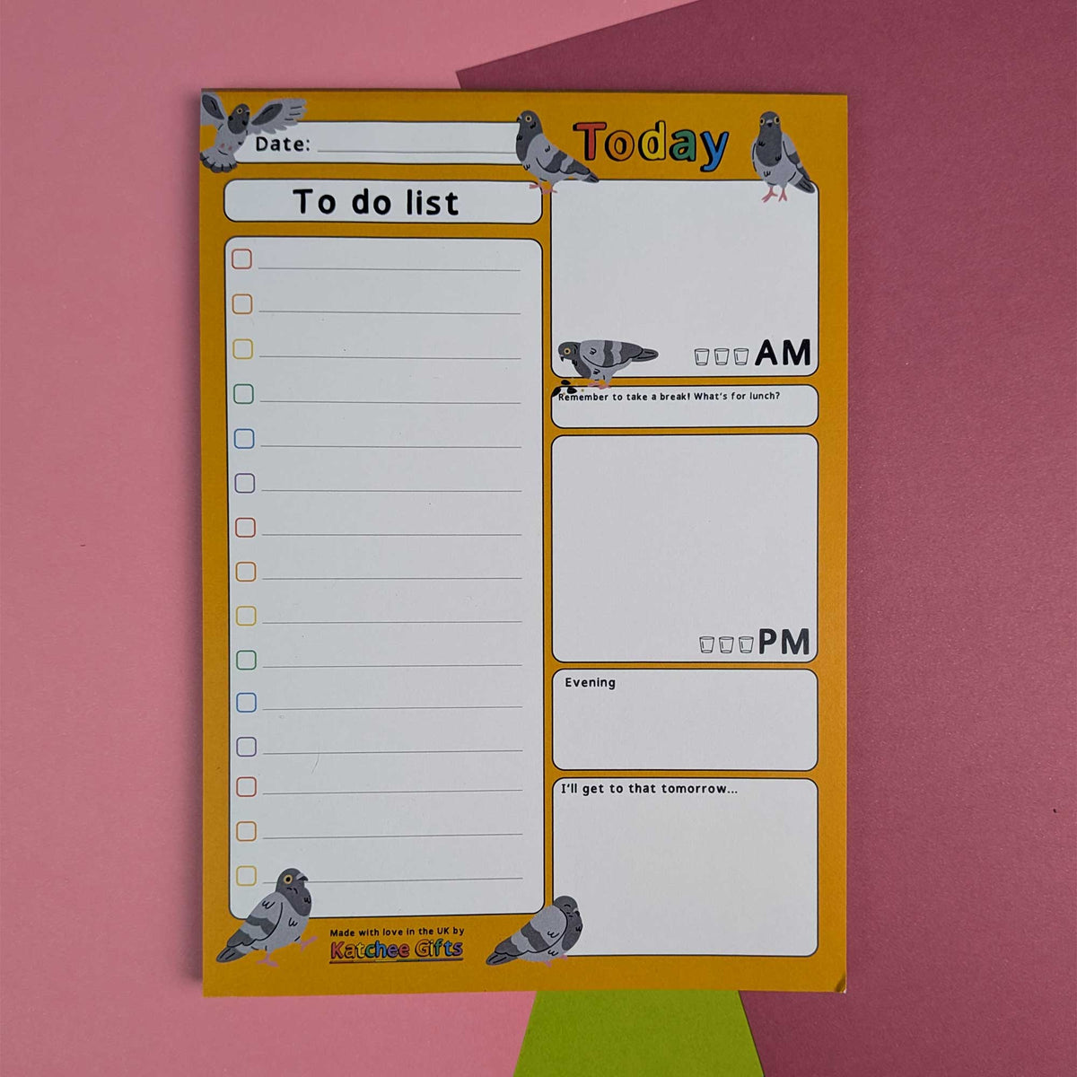 A daily to-do list notepad with pigeon illustrations, featuring a yellow border and white writing sections, photographed flat on a pink background with a green shape peeking from behind.